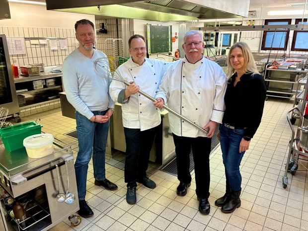 Vier Personen stehen in einer Großküche, zwei tragen Kochjacken und halten einen großen Schneebesen.