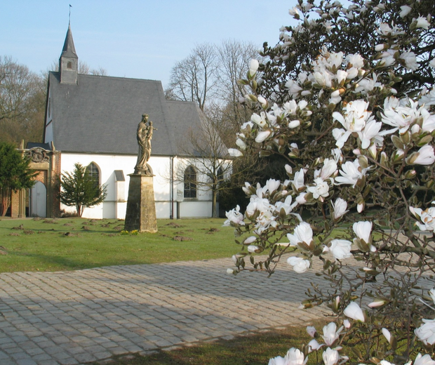 Das Bild zeigt die weiße Schlosskapelle, umgeben von Wiese und Bäumen. Im Vordergrund steht eine Statue. Bildquelle: LWL/Ritzenhoff