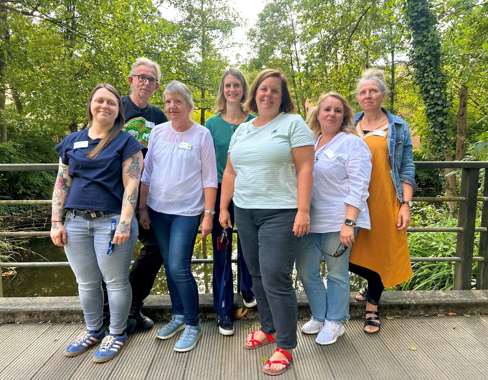 Eine Gruppe von sieben Personen steht im Freien auf einer Brücke und blickt in die Kamera. Im Hintergrund sind Bäume und ein Bach zu sehen.