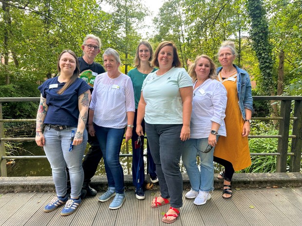 Eine Gruppe von sieben Personen steht im Freien auf einer Brücke und blickt in die Kamera. Im Hintergrund sind Bäume und ein Bach zu sehen.