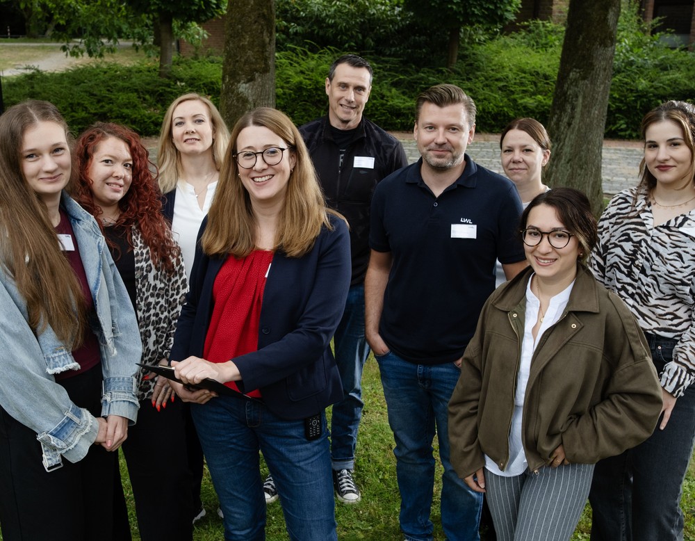 Gruppenfoto mit 9 Personen auf einer Wiese vor einer kleinen Mauer, Bäumen und Sträuchern