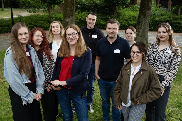Gruppenfoto mit 9 Personen auf einer Wiese vor einer kleinen Mauer, Bäumen und Sträuchern