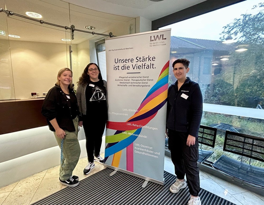 Drei Personen stehen in einem Eingangsbereich neben einem Roll-up mit der Aufschrift „Unsere Stärke ist die Vielfalt“. Das Plakat zeigt farbige Streifen und Logos des LWL. Im Hintergrund sind Glaswände, Stühle und ein Blick nach draußen zu sehen.