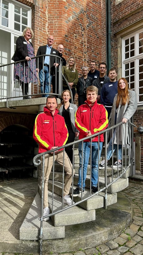 Eine Gruppe von Menschen steht auf einer Treppe im Freien und blickt in die Kamera, vorne tragen zwei Personen rote DLRG-Jacken.
