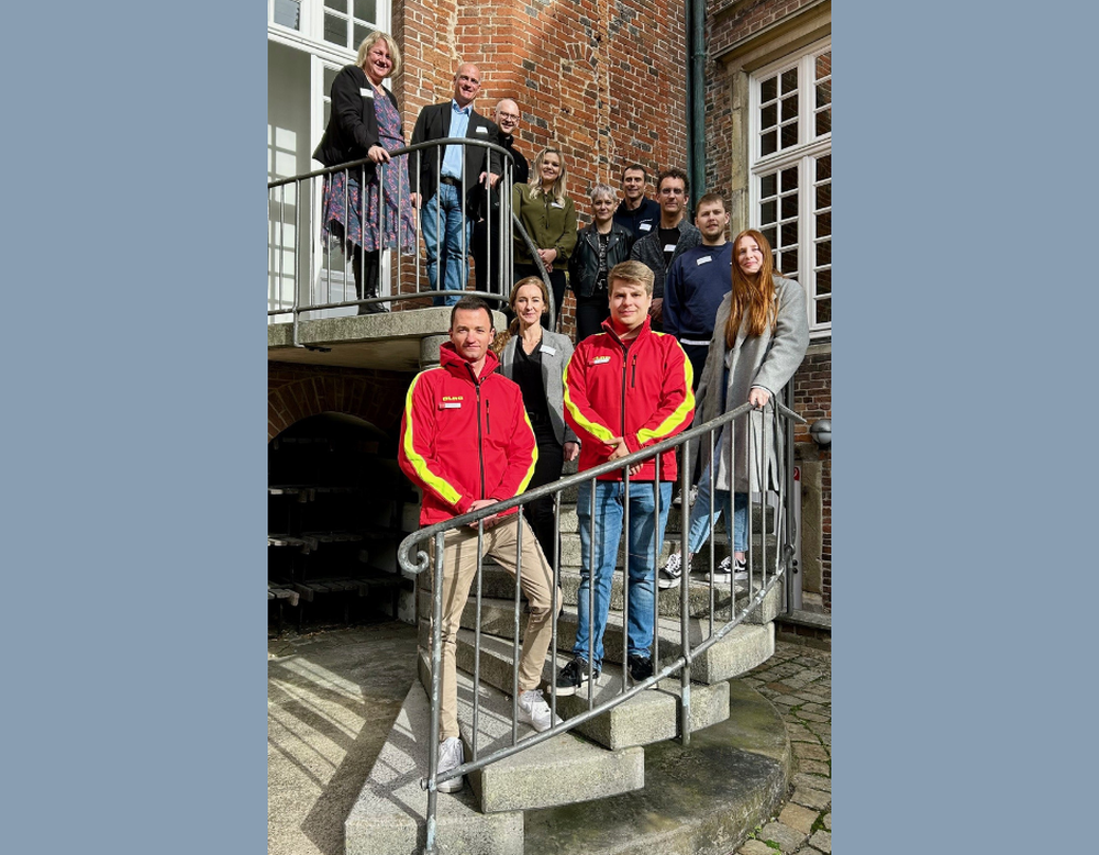 Eine Gruppe von Menschen steht auf einer Treppe im Freien und blickt in die Kamera, vorne tragen zwei Personen rote DLRG-Jacken.