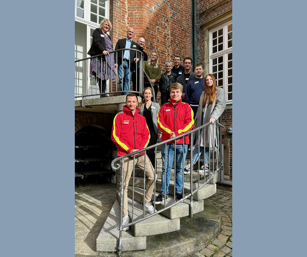 Eine Gruppe von Menschen steht auf einer Treppe im Freien und blickt in die Kamera, vorne tragen zwei Personen rote DLRG-Jacken.