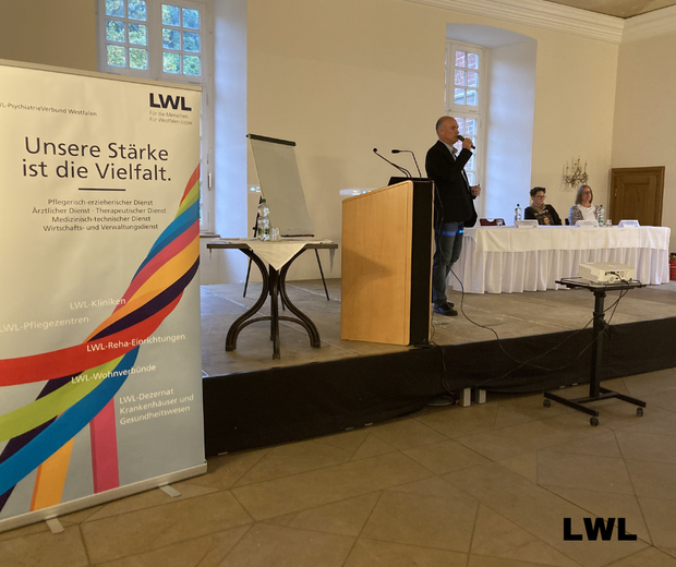 Ein Mann steht auf einer Vortragsbühne und spricht ins Mikrofon. Ein Mann steht auf einer Vortragsbühne und spricht ins Mikrofon.