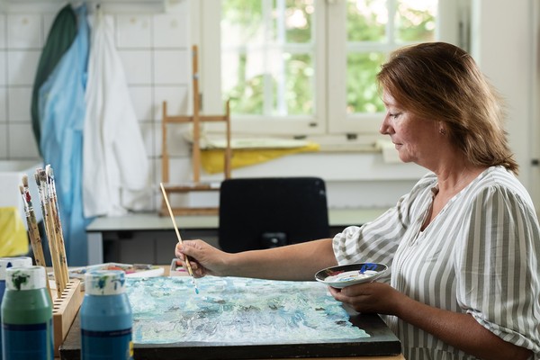 Eine Frau malt konzentriert an einem Bild in einem Atelier – auf dem Tisch stehen Pinsel und bunte Farbtuben.