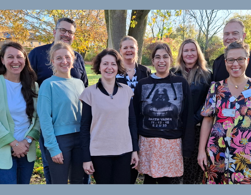 Eine Gruppe von neun Personen steht draußen auf einer Wiese und lächelt in die Kamera. Im Hintergrund sind herbstlich gefärbte Bäume zu sehen.