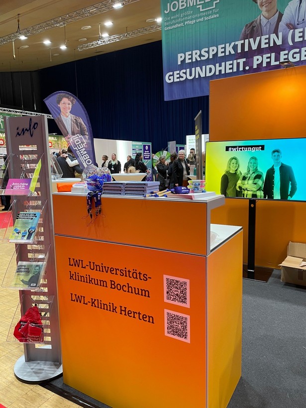 Blick auf einen Messestand des LWL mit Infomaterialien und einem Bildschirm, umgeben von Messebesucherinnen und -besuchern.