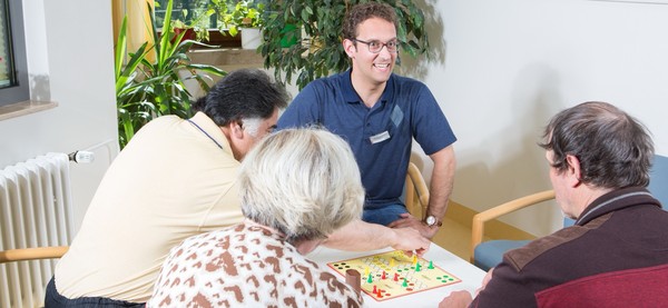 Ein Pfleger und drei Patienten sitzen in einem hellen Raum am Tisch und spielen das Spiel "Mensch ärger dich nicht" zusammen. LWL/Dirk Kaltenhäuser