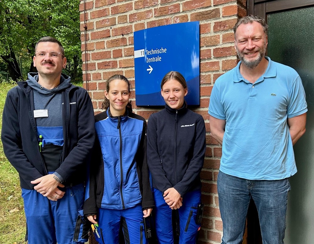 Vier Personen stehen lächelnd vor einem Backsteingebäude mit einem Schild „Technische Zentrale“. Zwei von ihnen tragen blaue Arbeitskleidung, die anderen Freizeitkleidung.