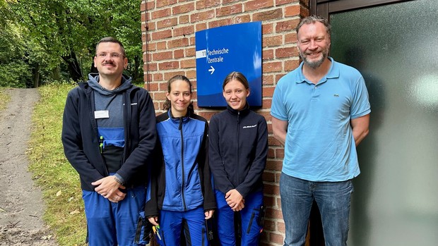 Vier Personen stehen lächelnd vor einem Backsteingebäude mit einem Schild „Technische Zentrale“. Zwei von ihnen tragen blaue Arbeitskleidung, die anderen Freizeitkleidung.