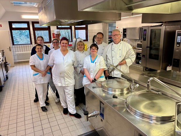 Neun Mitarbeitende stehen gemeinsam in einer Großküche vor Edelstahlgeräten und Arbeitsflächen.