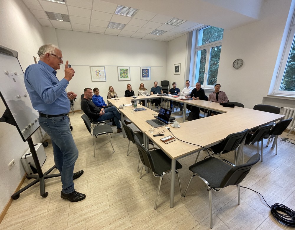 Schulungsraum mit Schulungsleiter vor einem FlipChart und 7 Teilnehmenden an Tischen sitzend.