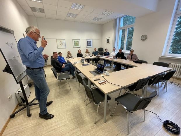 Schulungsraum mit Schulungsleiter vor einem FlipChart und 7 Teilnehmenden an Tischen sitzend.