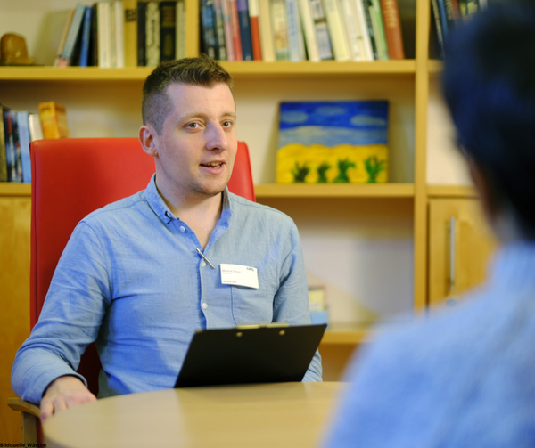 Ein Klinik-Mitarbeiter führt ein Gespräch mit einer Patientin im Vordergrund, im Hintergrund sind Bücherregale zu sehen. Bildquelle: LWL/Wäsche