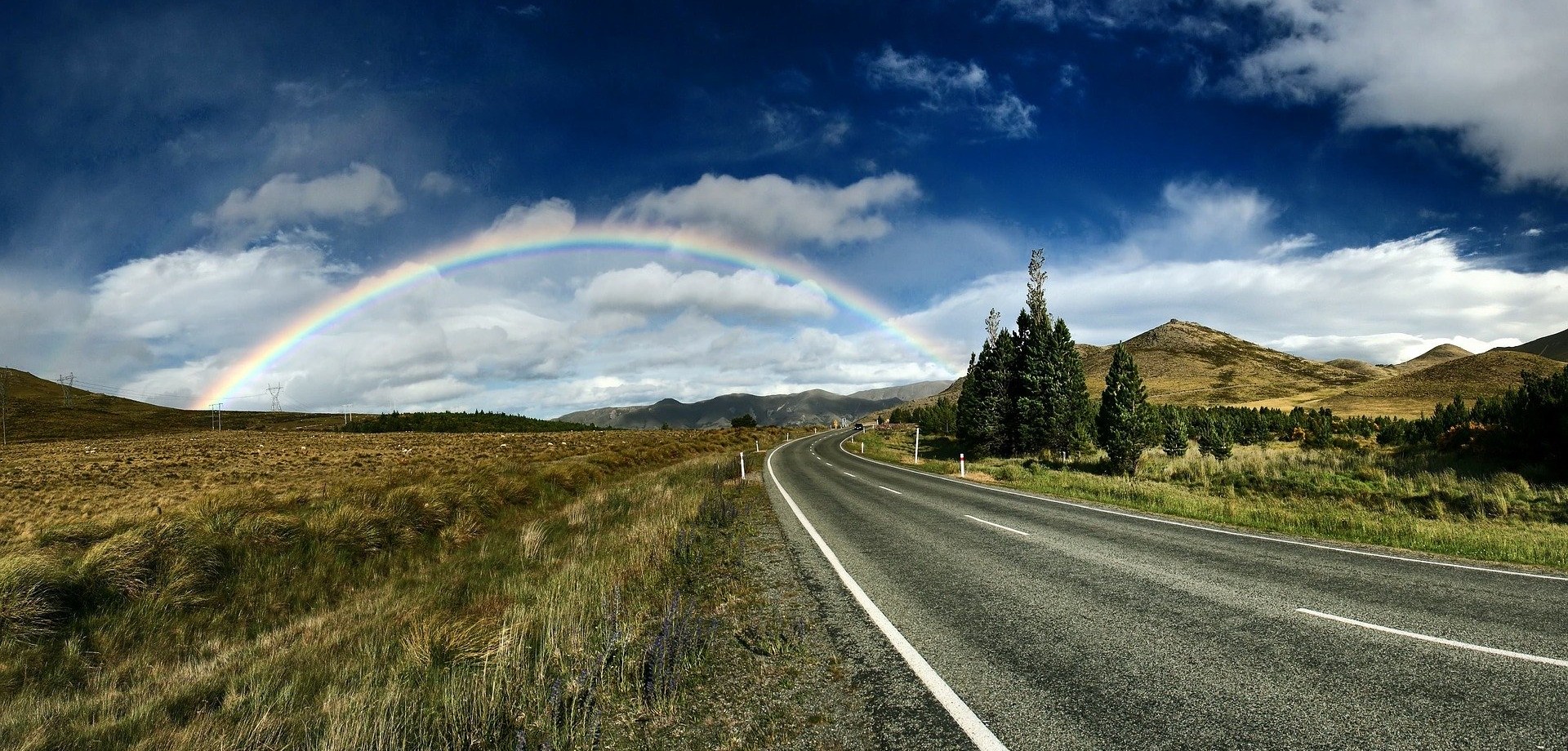 Durch eine karge Hügellandschaft führt eine langgezogene Straße und am Ende der Straße ist ein wunderschöner Regenbogen zu sehen