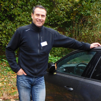 Eine Person steht im Freien neben einem Auto und lehnt mit einer Hand an der Autotür. Im Hintergrund sind herbstliche Büsche und Bäume zu sehen.