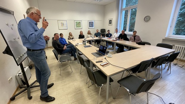 Schulungsraum mit Schulungsleiter vor einem FlipChart und 7 Teilnehmenden an Tischen sitzend.