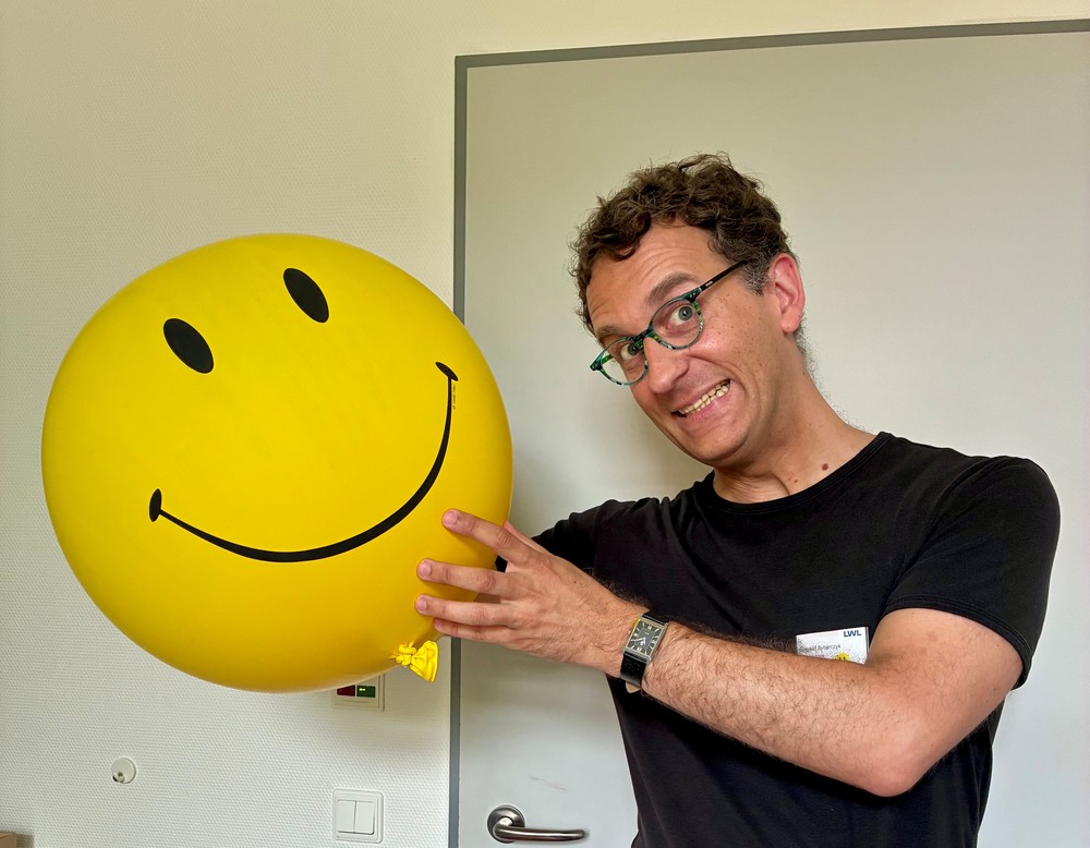 Ein Mann mit Brille und schwarzem T-Shirt hält einen großen gelben Luftballon mit aufgedrucktem Smiley in den Händen und lächelt in die Kamera.