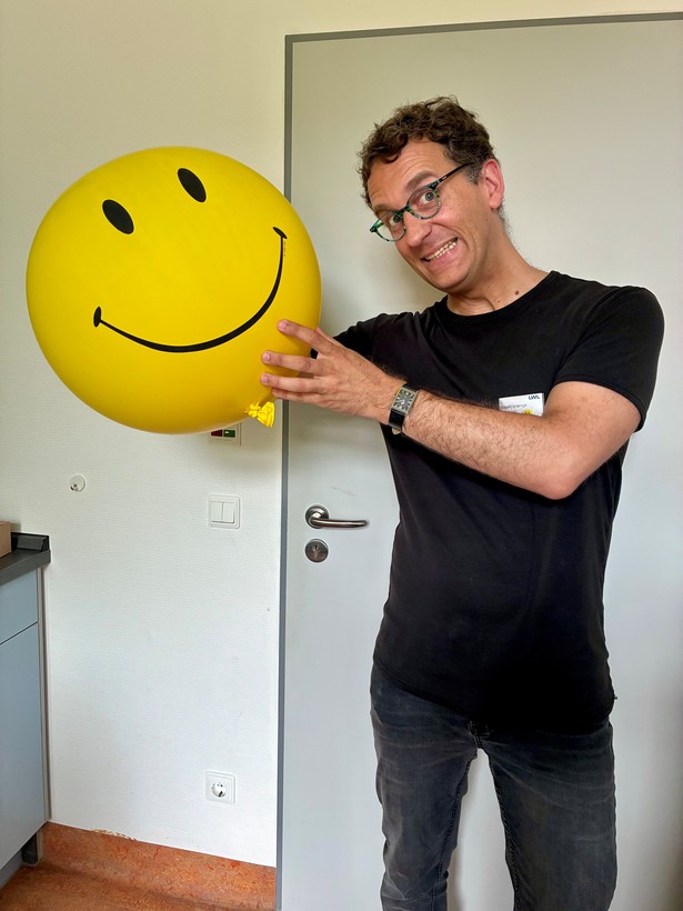 Ein Mann mit Brille und schwarzem T-Shirt hält einen großen gelben Luftballon mit aufgedrucktem Smiley in den Händen und lächelt in die Kamera.