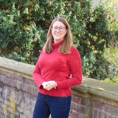 Eine Frau steht draußen vor einer niedrigen Steinmauer, dahinter dichte grüne Bäume.