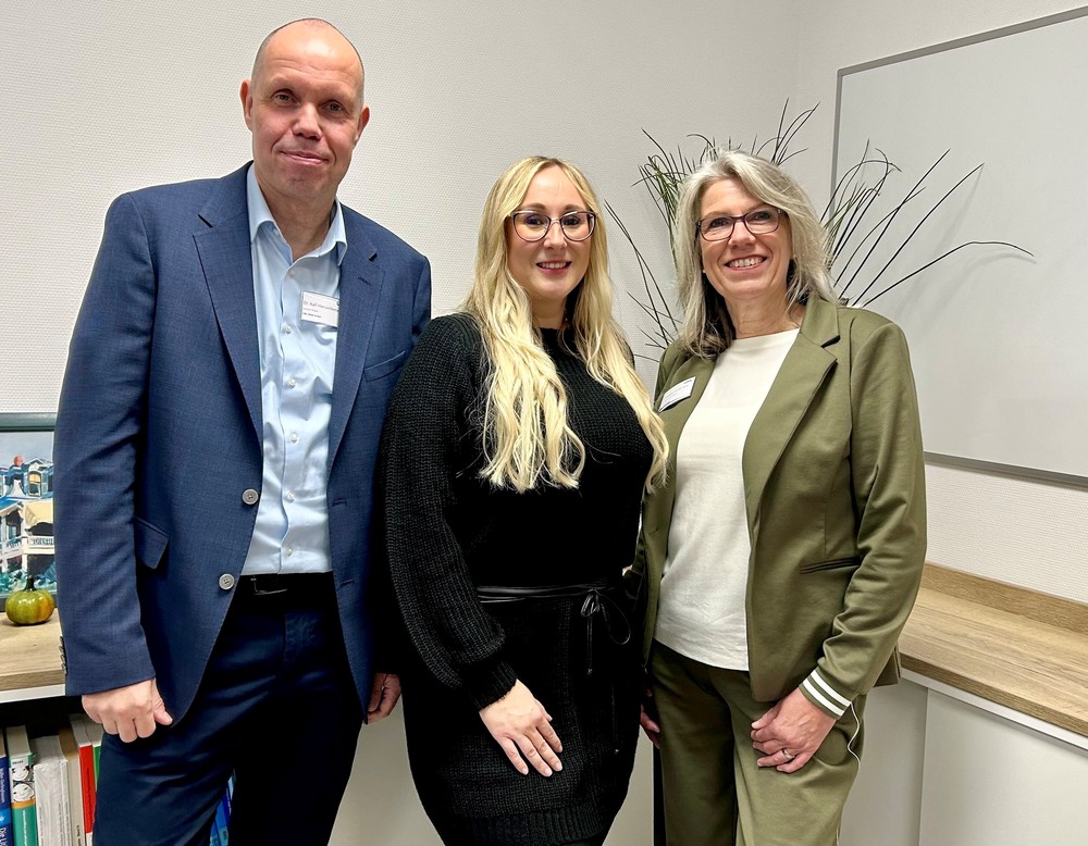 Drei Personen stehen in einem Büro nebeneinander und blicken in die Kamera. Hinter ihnen befinden sich ein Whiteboard, ein Regal mit Büchern und ein Bild auf einer Ablage.