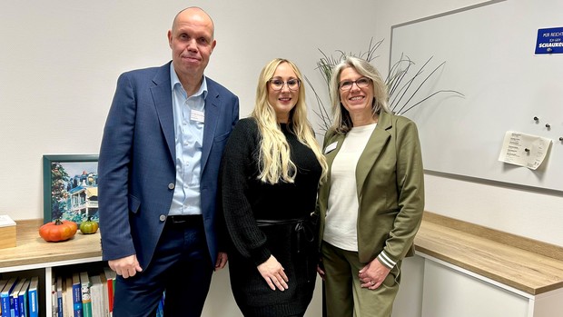 Drei Personen stehen in einem Büro nebeneinander und blicken in die Kamera. Hinter ihnen befinden sich ein Whiteboard, ein Regal mit Büchern und ein Bild auf einer Ablage.
