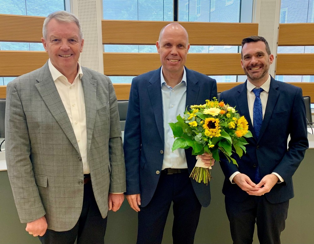 Drei Männer in Anzügen stehen nebeneinander und lächeln in die Kamera. Der Mann in der Mitte hält einen Strauß gelber Blumen mit Sonnenblumen in der Hand.