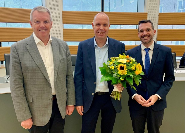 Drei Männer in Anzügen stehen nebeneinander und lächeln in die Kamera. Der Mann in der Mitte hält einen Strauß gelber Blumen mit Sonnenblumen in der Hand.