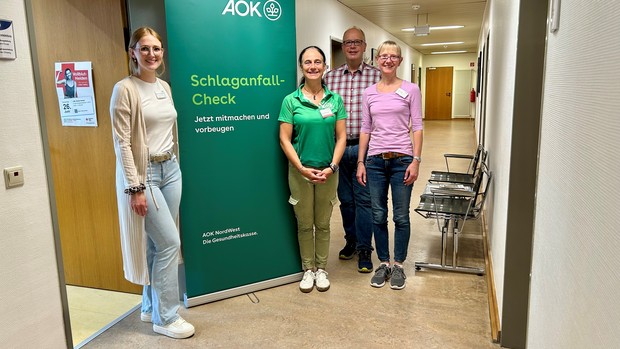 Vier Personen stehen neben einem grünen Informationsbanner der AOK mit der Aufschrift „Schlaganfall-Check – Jetzt mitmachen und vorbeugen“. Die Gruppe besteht aus Mitarbeitenden der AOK sowie der LWL-Klinik Herten..