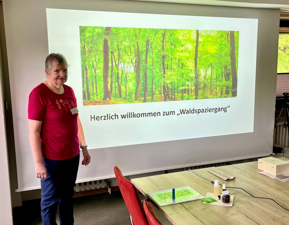 Eine Frau steht neben einer Leinwand, auf der eine Waldszene mit dem Text "Herzlich willkommen zum Waldspaziergang" projiziert wird.