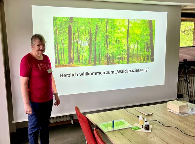 Eine Frau steht neben einer Leinwand, auf der eine Waldszene mit dem Text "Herzlich willkommen zum Waldspaziergang" projiziert wird. Eine Frau steht neben einer Leinwand, auf der eine Waldszene mit dem Text "Herzlich willkommen zum Waldspaziergang" projiziert wird.