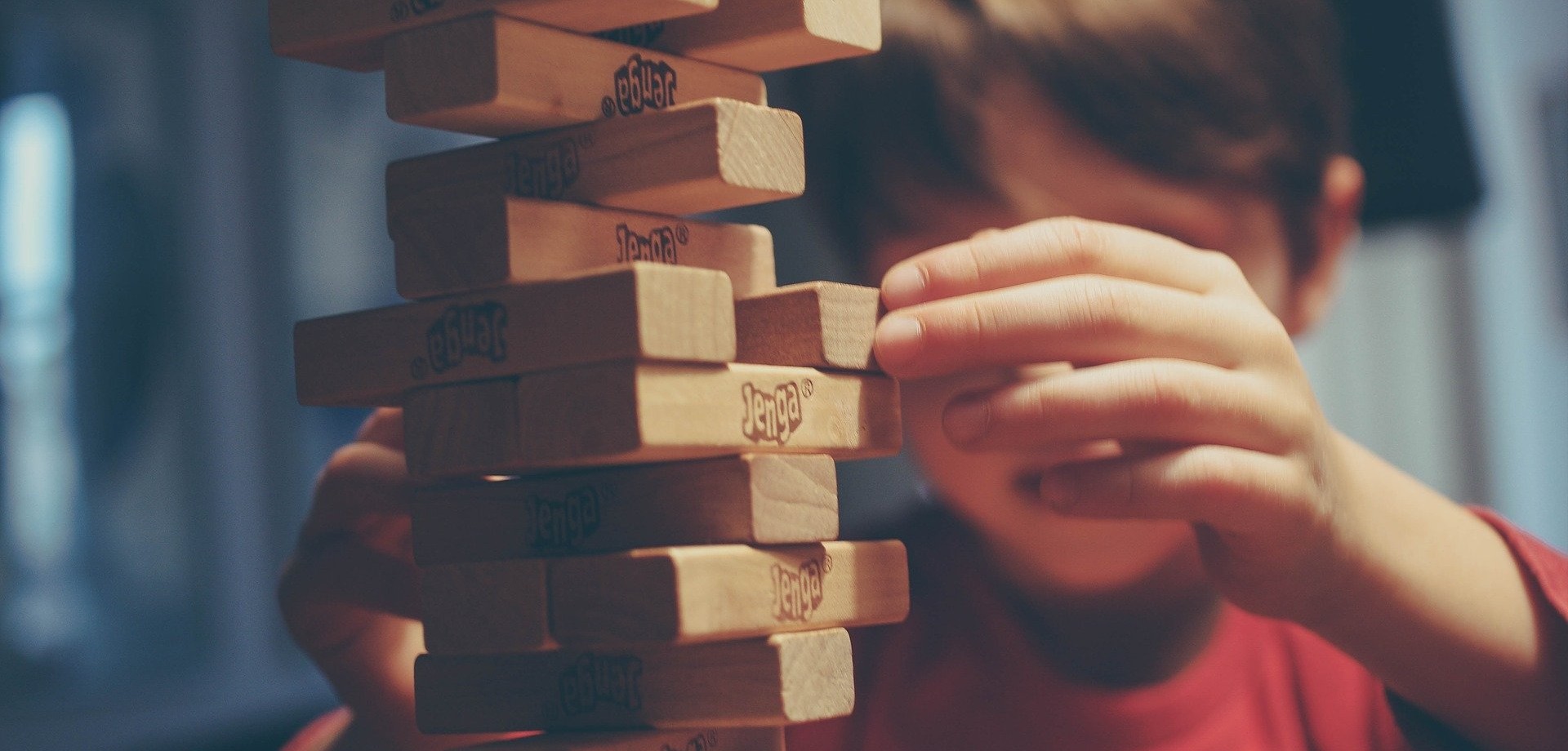 Ein kleiner Junge ist bei dem Spiel Jenga sehr weit gekommen und zieht vorsichtig einen weiteren Stab aus dem schon sehr wackeligen Turm