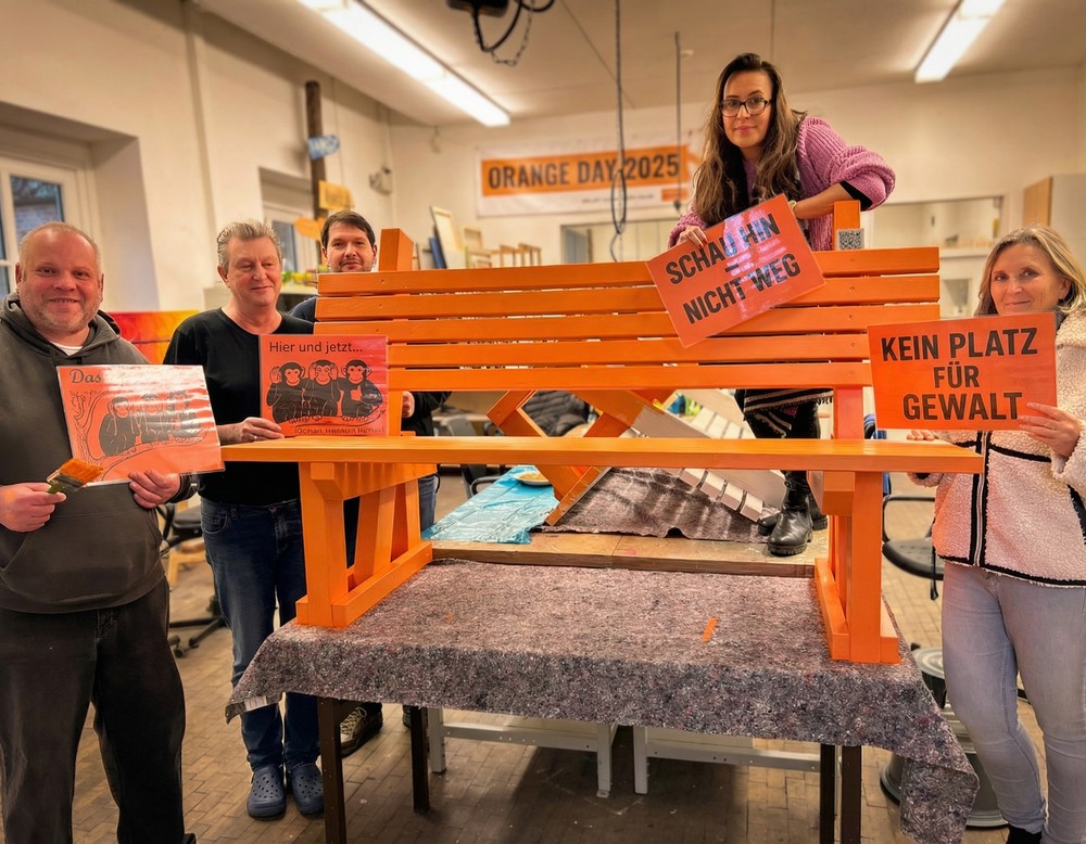 Fünf Personen stehen in einer Werkstatt um eine orange Bank und halten Schilder gegen Gewalt hoch.