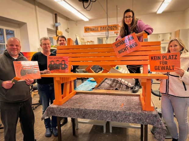 Fünf Personen stehen in einer Werkstatt um eine orange Bank und halten Schilder gegen Gewalt hoch.