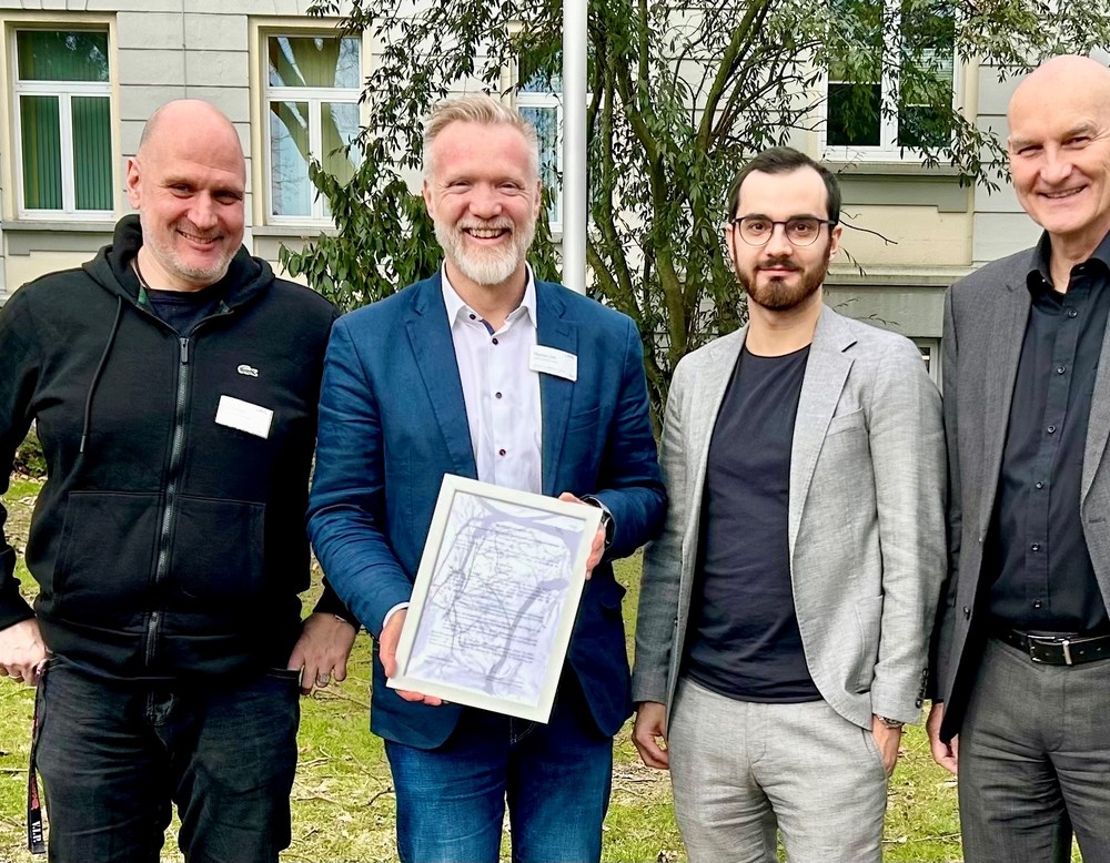 Vier Männer stehen vor einem Gebäude. Einer davom hält ein Zertifikat in den Händen.