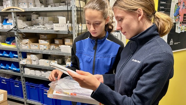 Zwei Personen in blauer Arbeitskleidung stehen in einer Werkstatt vor Regalen mit technischen Ersatzteilen. Sie betrachten gemeinsam eine Lampe oder ein Bauteil und besprechen offenbar die nächsten Arbeitsschritte.
