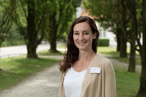 Eine Frau mit Namensschild steht auf einem von Bäumen gesäumten Weg im Freien und lächelt in die Kamera.