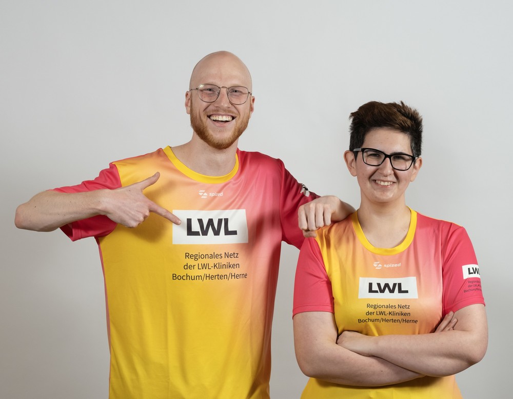 Zwei Personen tragen farbige LWL-Sportshirts und stehen vor neutralem Hintergrund