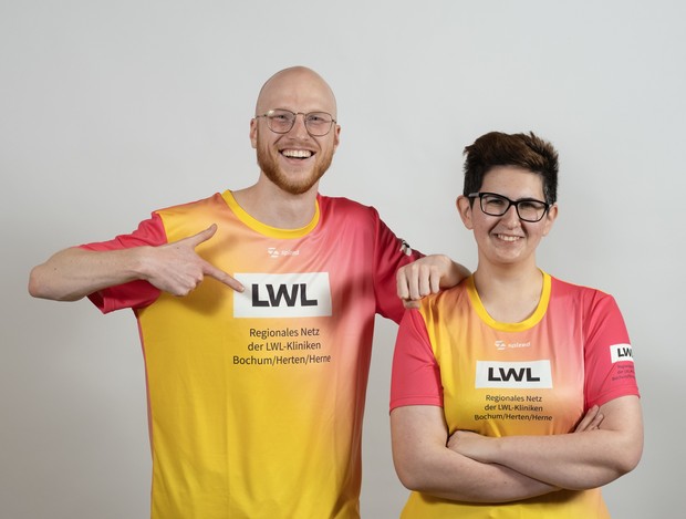 Zwei Personen tragen farbige LWL-Sportshirts und stehen vor neutralem Hintergrund