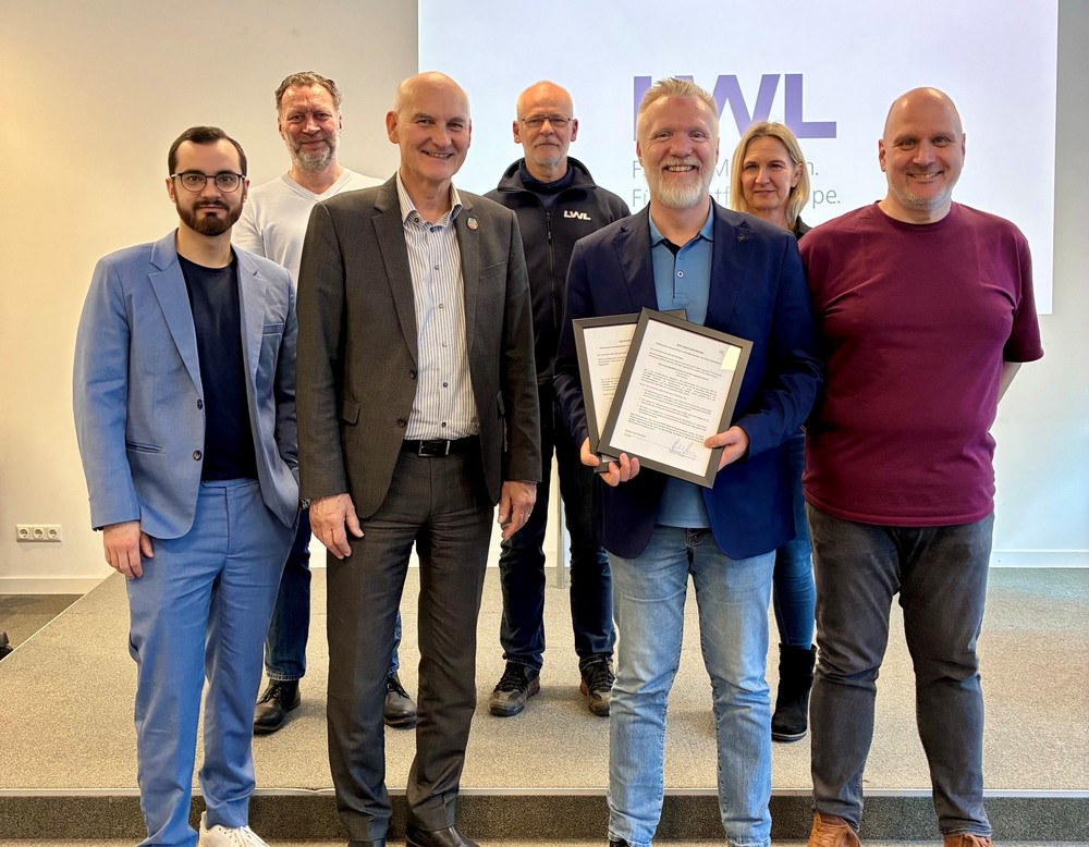 Gruppe von sieben Personen steht in einem Konferenzraum vor einem LWL-Hintergrund; eine Person hält zwei gerahmte Urkunden.