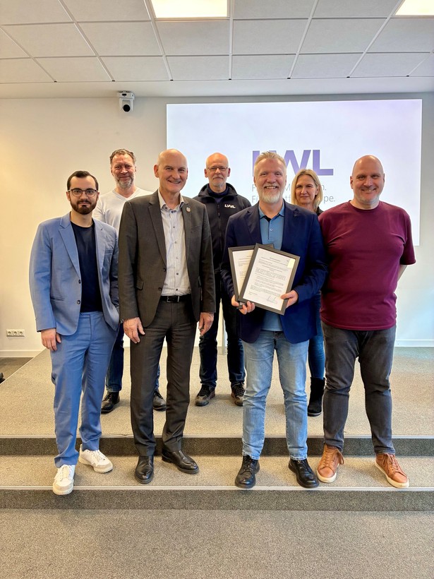 Gruppe von sieben Personen steht in einem Konferenzraum vor einem LWL-Hintergrund; eine Person hält zwei gerahmte Urkunden.