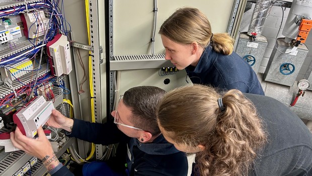 Drei Personen arbeiten gemeinsam an einem geöffneten Schaltschrank. Eine Person zeigt den beiden anderen ein technisches Bauteil, während viele bunte Kabel sichtbar sind.