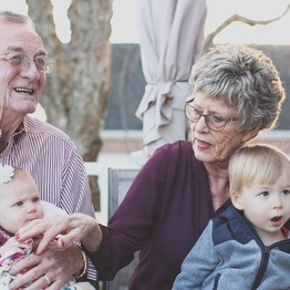 Oma und Opa sitzen jeweils auf einem Stuhl und haben eines ihrer Enkelkinder auf dem Arm