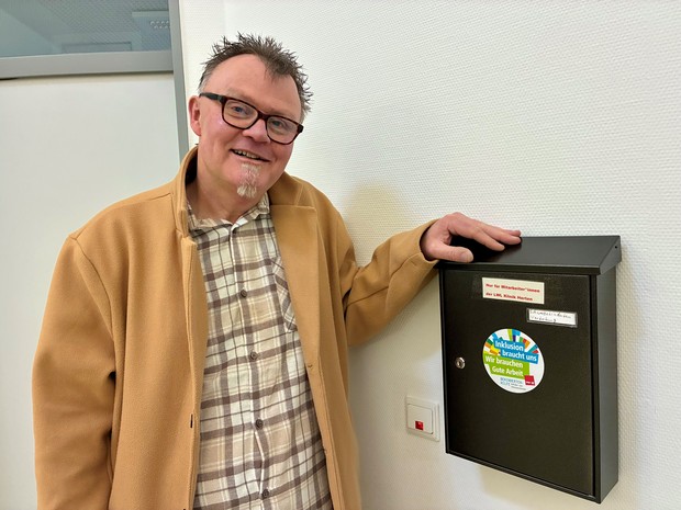 Ein Mann mit Brille steht neben einem schwarzen Briefkasten mit Aufkleber zum Thema Inklusion.